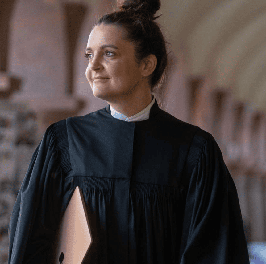 Porträt von Online-Pfarrerin Jennifer Scherf. Sie hält einen LapTop unter dem rechen Arm und trägt eine schwarzen Thala. Im Hintergrund ist eine verschwommene, helle Umgebung zu sehen.